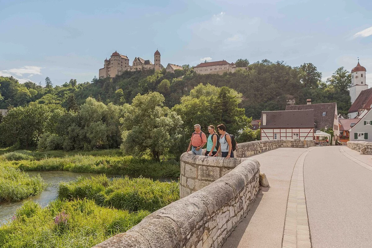 Drei Wanderer stehen auf der steinernen Brücke und blicken auf die Wörnitz. Links oben thront über dem Wald die Harburg.