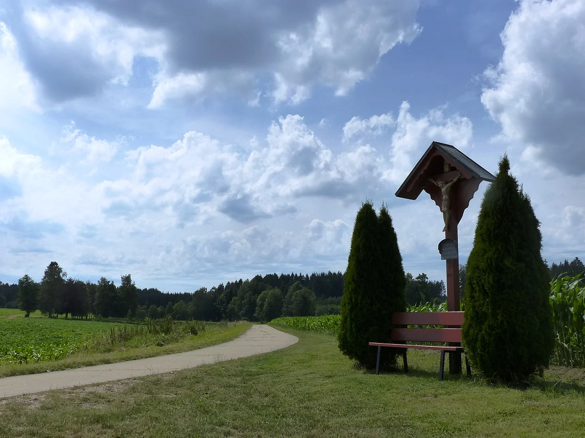 Blick auf ein idyllisches Fleckchen - ein Wegkreuz mit Holzbank davor - im Hintergrund sieht man den Wald.