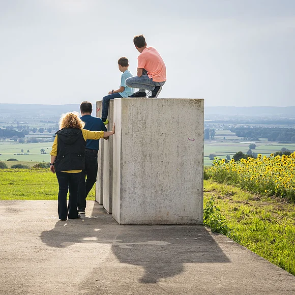 Die Kinder sitzen auf den Steinen der Zeitpyramide und die Eltern stehen daneben. Gemeinsam blicken sie in das Ries und genießen den tollen Ausblick.