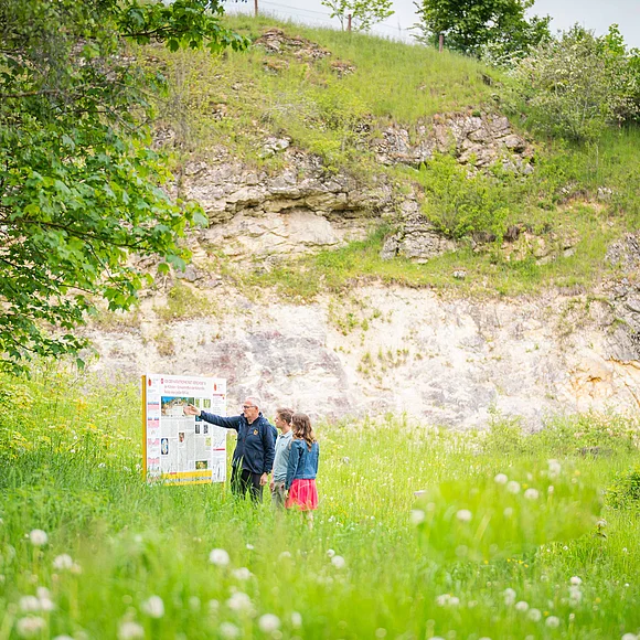 Drei Personen stehen auf einer Wiese mit hohem Gras vor einer Informationstafel. Im Hintergrund ist eine kleine Felswand zu sehen.