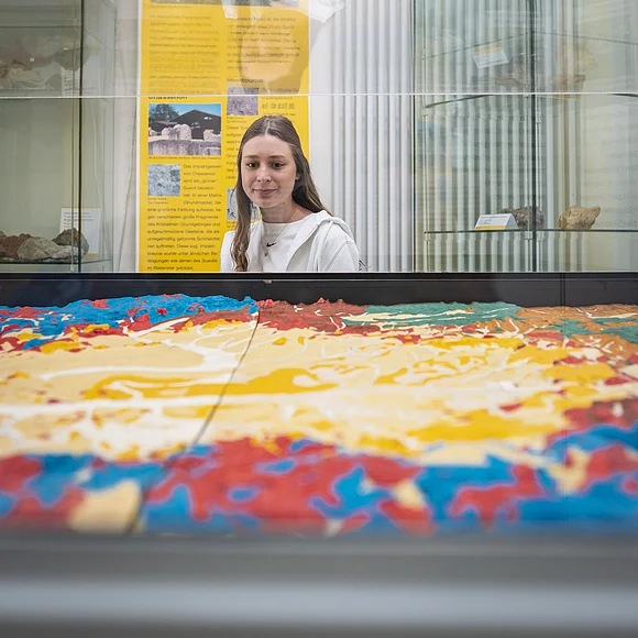 Ein Mädchen betrachtet ein bunt eingefärbtes Modell einer Landschaft in einer Vitrine. Im Hintergrund ist eine gelbe Informationstafel zu sehen.