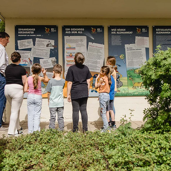 Ein Mann und eine Gruppe Kinder betrachtet Informationstafeln an einer Hauswand.