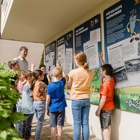 Ein Mann und eine Gruppe Kinder betrachtet Informationstafeln an einer Hauswand.