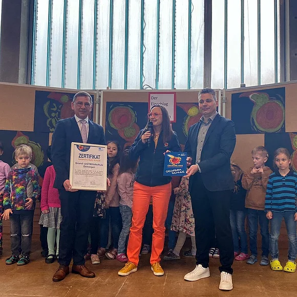 Heike Burkhardt überreicht Wilhelm Rehklau und Andreas Langer die Urkunde der Geopark Ries Schule Deiningen. Dahinter stehen einige Kinder der Schule und gemalte Bilder des Meteoriteneinschlags.