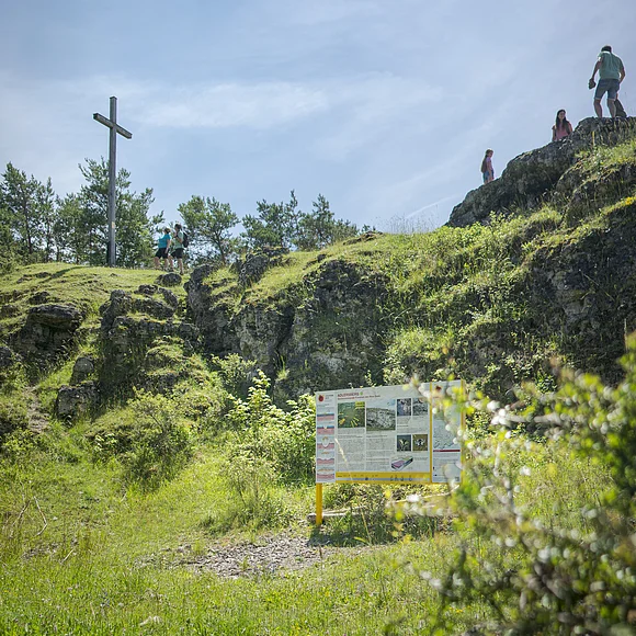 Geopark-Shooting 2021 Auf einer mit Gras bewachsenen Felsformation stehen ein Kreuz und einige Personen. Davor steht eine Informationstafel.