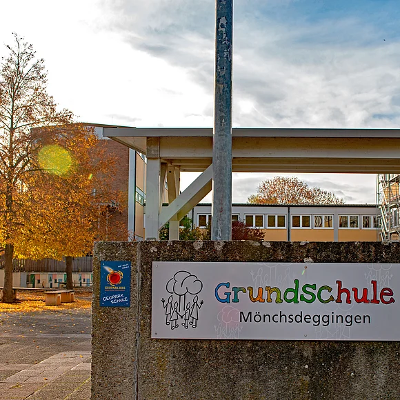 An einer niedrigen Betonmauer ist ein Schild mit der Aufschrift "Geopark Ries Schule" und ein großes Schild mit der Aufschrift "Grundschule Mönchsdeggingen" angebracht. Im Hintergrund sind mehrere Gebäude zu sehen.
