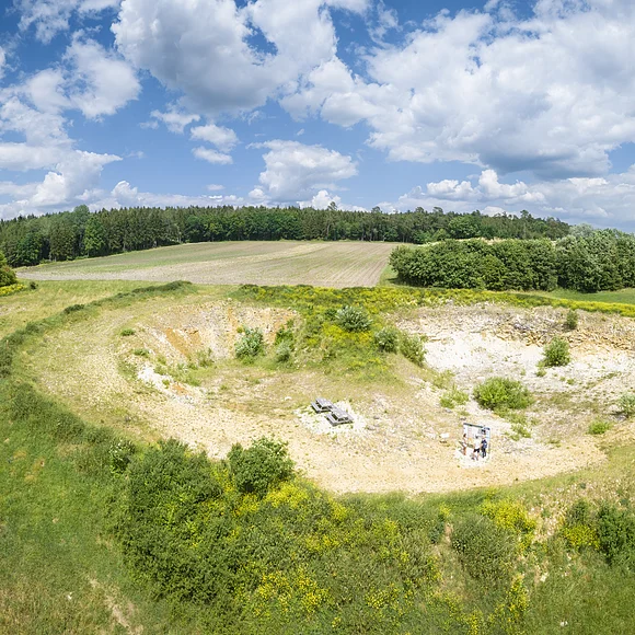 In einer flachen Grube stehen vereinzelt Büsche, während die Hänge aus bloßem Gestein bestehen. Um die Grube herum befinden sich Wälder, Äcker und Wiesen.