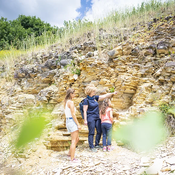 Eine Geopark Ries Führerin zeigt einer Frau und einem Mädchen eine Felswand aus plattigem Kalkgestein. Der Boden ist ebenfalls mit Bruchstücken von Kalkgesteinsplatten bedeckt.