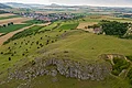 Hinter einem Hügel mit Wiese und felsiger Stirn erstreckt sich eine weite Landschaft mit Siedlungen, Äckern, Wiesen und Bäumen.