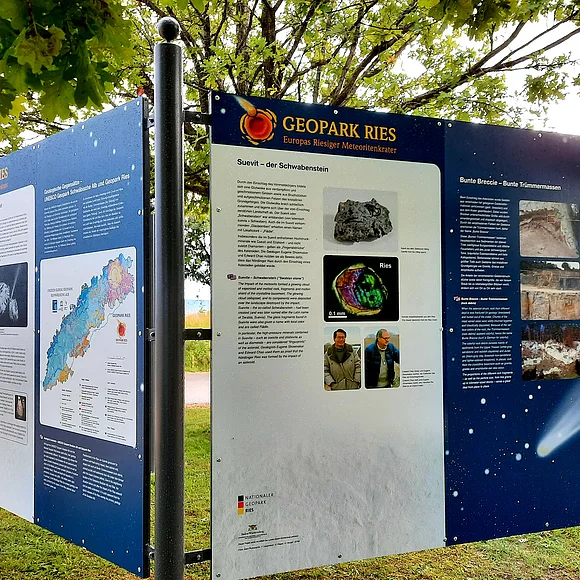 Unter Bäumen stehen zwei Informationstafeln über den Geopark Ries mit einer geologischen Karte und Informationen zum Schwabenstein Suevit.