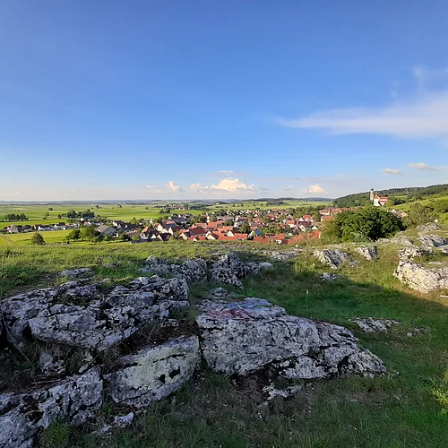 Ausblick vom Geotop Kühstein, Mönchsdeggingen Von einem Hügel mit felsiger Kuppe aus erstreckt sich unter blauem Himmel der Blick in eine weite Landschaft mit einer Siedlung, Äckern, Wiesen und Bäumen.