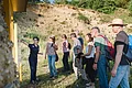 Eine Frau zeigt für eine Gruppe Personen auf eine Informationstafel. Im Hintergrund ist ein felsiger Hanganriss zu sehen.