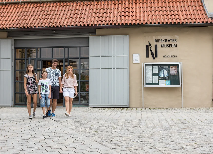Eine Familie geht auf dem gepflasterten Platz vor einer ehemaligen Scheune. An deren Wand befindet sich die Aufschrift "Rieskrater Museum Nördlingen".