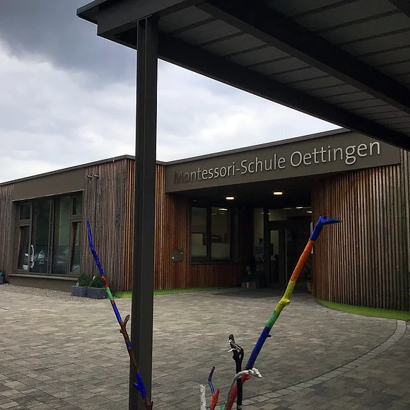 Montessori-Schule Oettingen Seitlicher Blick auf den Eingang der Montessori-Schule Oettingen. Im Vordergrund ein bunt bemalter Ast.