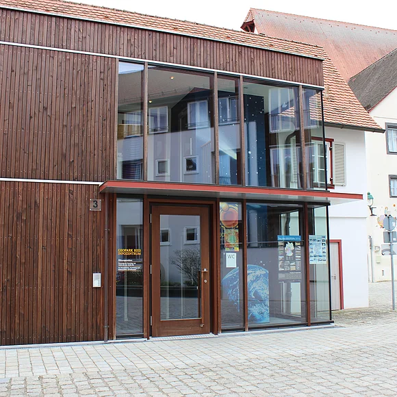 An einem gepflasterten Platz steht ein zweistöckiges Gebäude mit brauner Holzfassade und großen Fenster.