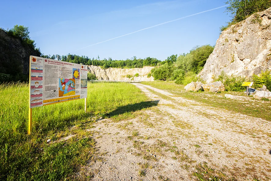 Neben einem Schotterweg, der in einen ehemaligen Steinbruch führt, steht eine große Informationstafel.