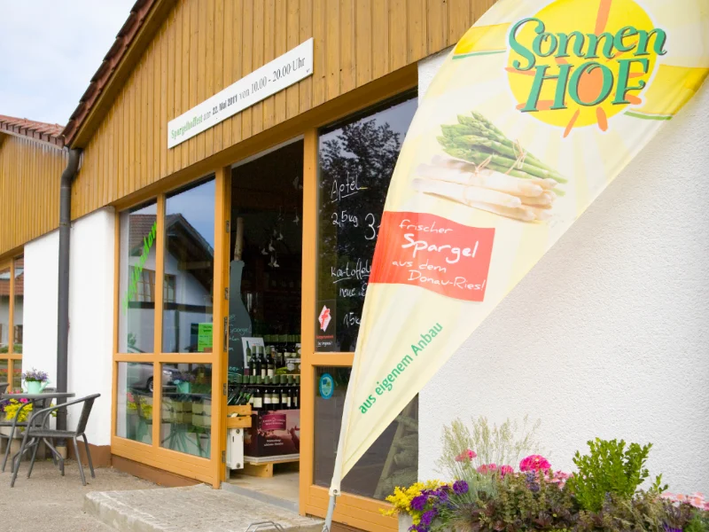Sonnenhof Durch eine geöffnete Glastür ist eine Palette mit Weinflaschen zu sehen. Neben der Tür steht eine Fahne mit der Aufschrift "Sonnenhof" und "frischer Spargel aus dem Donau-Ries - aus eigenem Anbau".