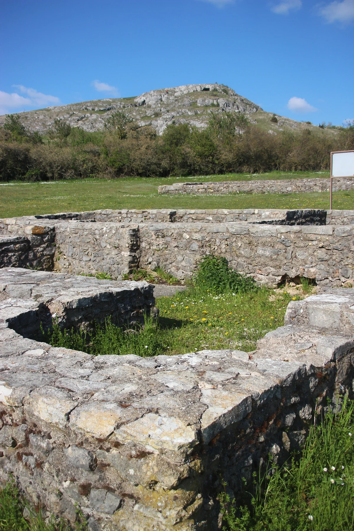 Auf einer Wiese befinden sich die Grundmauern eines Gebäudes. Im Hintergrund ist ein felsiger Hügel zu sehen.