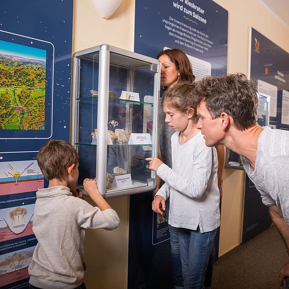 Eine Familie betrachtet eine kleine Glasvitrine mit Gesteinsbruchstücken. An der Wand befinden sich dunkelblaue Informationstafeln über den Meteoriteneinschlag im Ries.