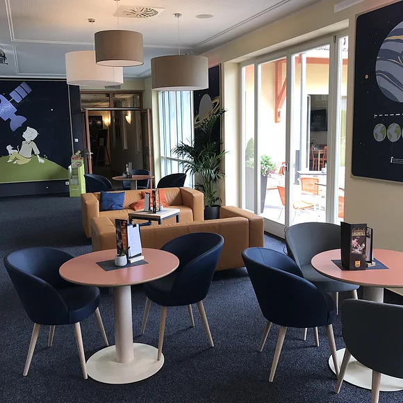 In einem Raum stehen verschiedene Sitzgelegenheiten mit kleinen Tischen. An der Wand hängen großformatige Zeichnungen von Planeten.