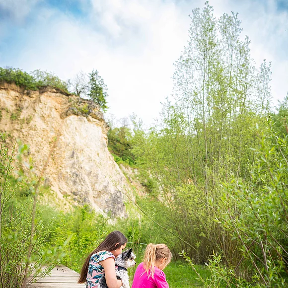 Zwei Mächen sitzen mit einem Hund auf einem Holzsteg über einen kleinen Teich. Im Hintergrund ist eine steil aufragende Felswand erkennbar.
