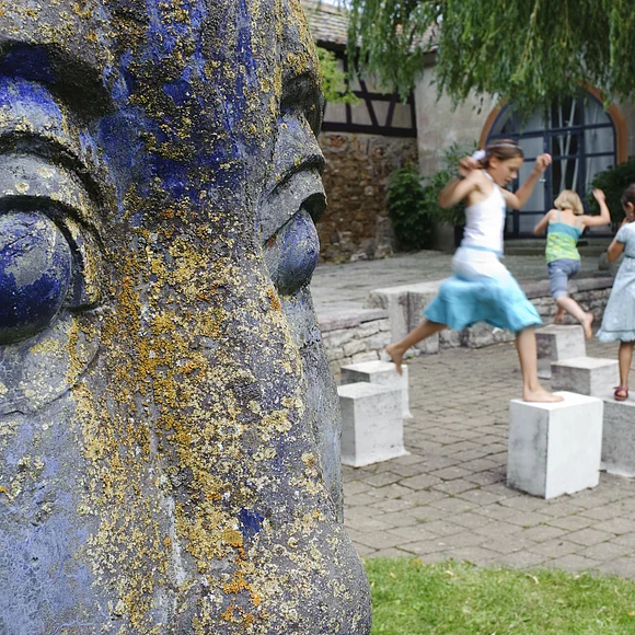 Rechts im Bild die Nahaufnahme des Gesichts einer blauen Statue. Dahinter stehen Steinwürfel in einem Hof. Drei junge Mädchen hüpfen von einem Würfel zum anderen.