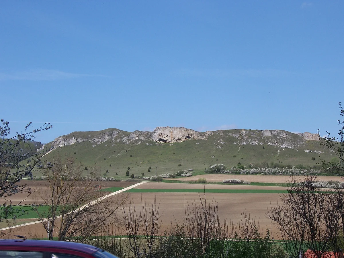 Hinter einem großen Acker erhebt sich ein langgezogener Hügel mit grasbewachsenen Hängen und felsiger Kuppe.