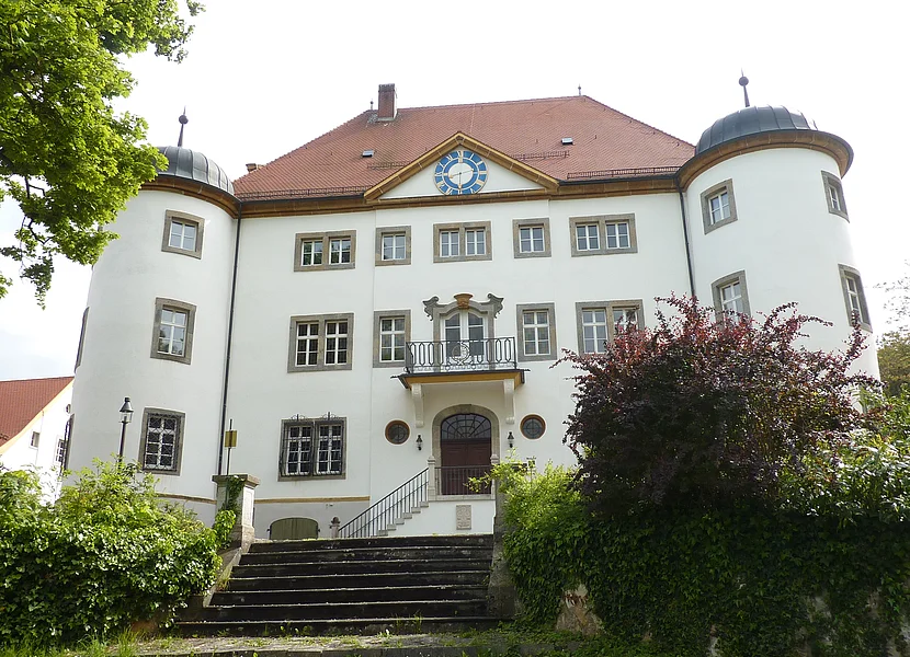 Castello Schloss Reimlingen Ein dreistöckiges Gebäude mit weißer Fassade hat an den Seiten runde Türme und am Giebel eine blaue Uhr.