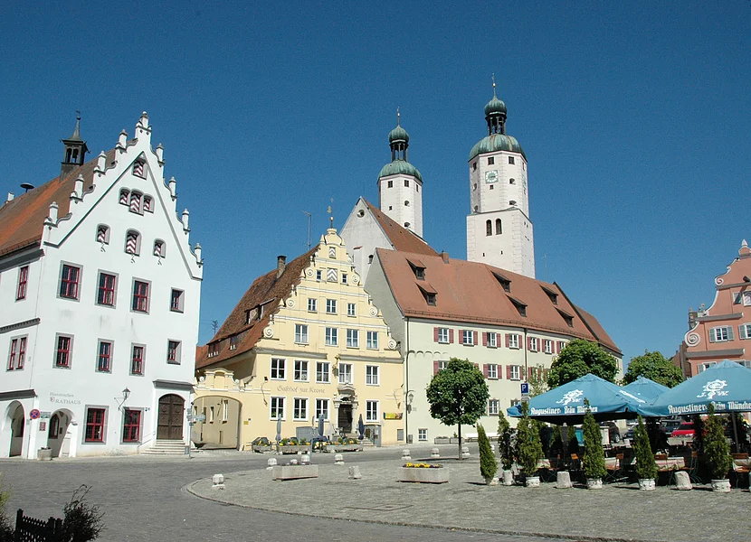 Piazza del mercato (Wemding) In einer Innenstadt stehen verschiedene historische Gebäude, hinter denen sich zwei Kirchtürme erheben. Am linken Bildrand stehen blaue Sonnenschirme.