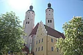 Zwei weiße Kirchtürme mit grünlichen Dächern ragen in den Himmel. Davor ist ein gelbes Haus und Bäume zu sehen.