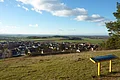 Von einem Hügel mit einer Panoramatafel aus erstreckt sich der Blick über eine Siedlung hinweg in eine weite Landschaft mit Äckern und Wald.