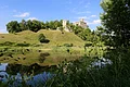 Auf einem Hügel mit Bäumen steht eine Burgruine, die sich in der ruhigen Wasserfläche im Vordergrund spiegelt.