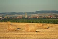 Auf einem abgeernteten Getreidefeld liegen Strohballen. Im Hintergrund lässt sich unscharf eine Siedlung mit Kirchturm und ein Hügelrücken erkennen.