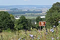 Hinter Bäumen erstreckt sich eine weite kesselförmige Landschaft mit Wald, Äckern und Siedlungen. Im Vordergrund ist ein braunes Schild mit dem Logo des Geopark Ries zu sehen.
