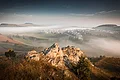 Von einem Hügel aus erstreckt sich der Blick in eine weite Landschaft mit Hügeln, einer Siedlung und großen Nebelschwaden. Im Vordergrund ist ein Felsen zu sehen.