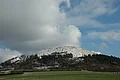 Ein Hügel ist an der Kuppe von Schnee bedeckt und im unteren Bereich mit Bäumen bewachsen.
