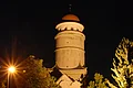 Über einer Straße erhebt sich ein beleuchtetes Stadttor mit rundem Turm. Der Himmel ist dunkel.