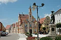 Neben einer Straße steht ein Haus mit weiß-oranger Fassade und kleinem Turm. Im Vordergrund ist ein Denkmal mit einer Kupferfigur auf einem Sockel zu sehen.