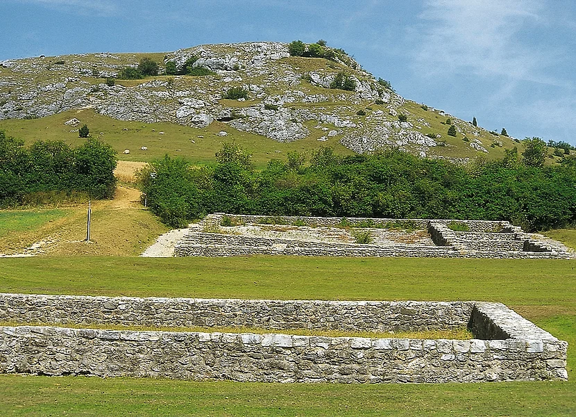Villa rustica (podere romano) a Holheim, II-III sec. d.C. In einer Wiese befinden sich Mauerfundamente zweier Gebäude. Hinter einer Buschreihe erhebt sich ein felsiger Hügel.