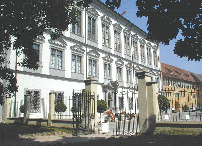 Residenza di Oettingen Ein dreistöckiges Schloss hat eine weiße Fassade und Fenster mit grauem Gesims. An der Seite schließt sich ein schmiedeeiserner Zaun an.