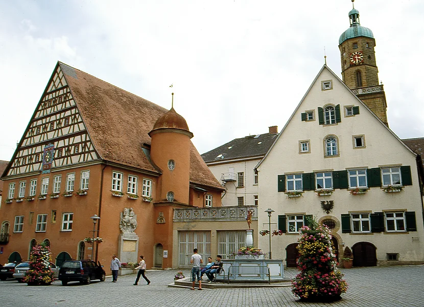 iazza del mercato (Bopfingen am Ipf) An einem gepflasterten Platz befinden sich ein rotes Gebäude mit Fachwerk am Giebel und ein cremefarbenes Gebäude mit Fensterläden, hinter dem sich ein Kirchturm erhebt.