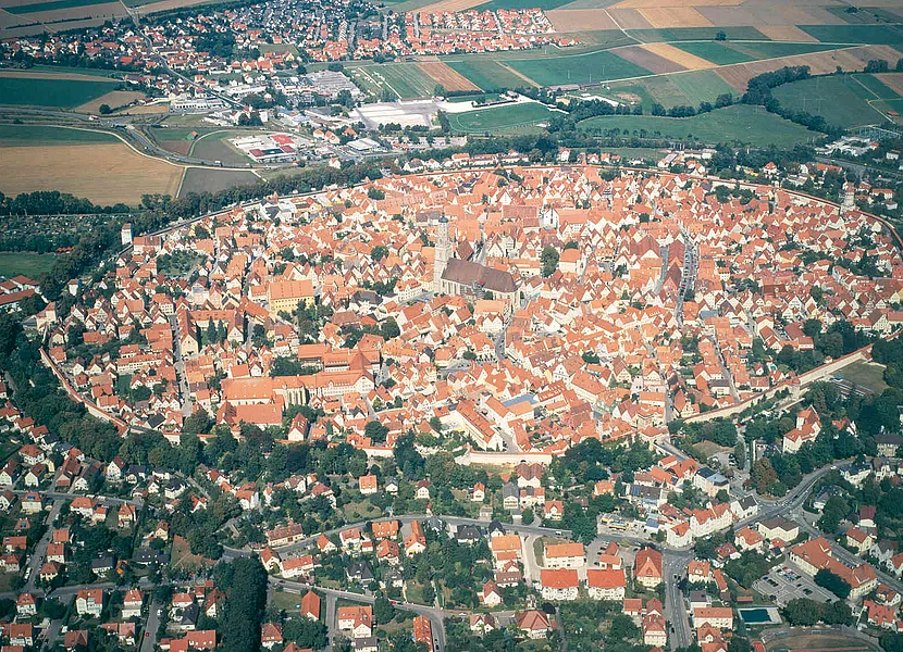 L’antica “libera città imperiale” di Nördlingen con la cinta muraria interamente conservata e percorribile Das Luftbild zeigt die kreisrunde Altstadt von Nördlingen mit dem Kirchturm in der Mitte. Außerhalb der Stadtmauer schließen sich weitere Siedlungen an.