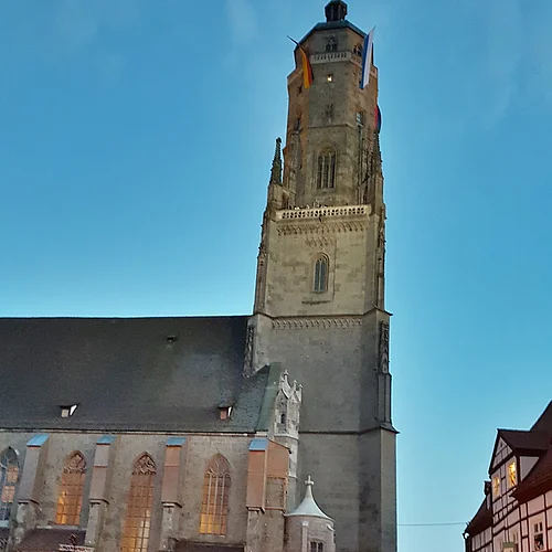 Auf dem Platz vor einer Kirche mit hohem Turm befinden sich viele Menschen, teils in mittelalterlicher Kleidung und mit Fackeln.