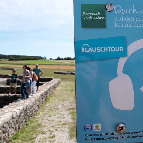 Lauschtour Rieskrater-Tour Ein kleines Schild weist auf die Lauschtour 5 "Durch den Rieskrater" hin. Im Hintergrund sind Personen zu sehen, die auf den Mauerfundamenten eines Gebäudes laufen.