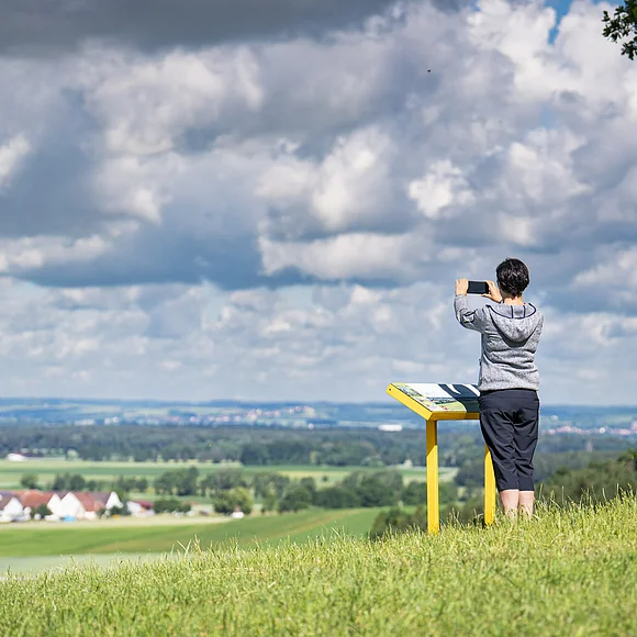 Eine Frau steht hinter einer Panoramatafel auf einer Wiese und macht ein Foto von der weiten Landschaft mit Siedlungen, Wald und Wiesen.