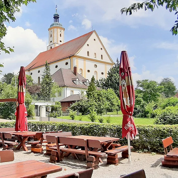 Im Vordergrund sieht man einen Teil des Biergartens mit seinen Tischen und Bänken. Im Hintergrund sieht man Wallfahrtsbasilika Maria Brünnlein.