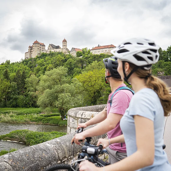 Zwei Personen mit Fahrrädern und Helmen stehen auf einer Brücke und blicken auf eine Burg auf einem Hügel.