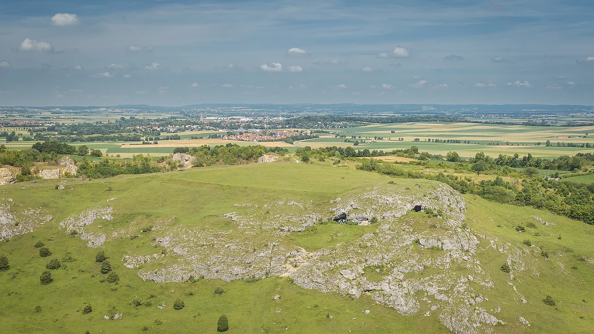 Luftbildaufnahme vom Riegelberg mit den Ofnethöhlen Hügelige Landschaft mit grünen Wiesen und Felsen im Vordergrund, Felder und ein Dorf im Hintergrund.