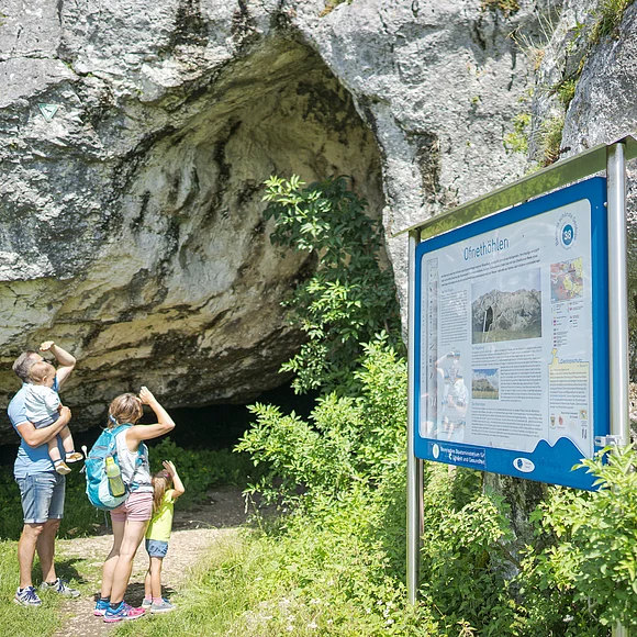 Familie an den Ofnethöhlen Eine Familie steht vor einer Höhle und betrachtet ein Informationsschild mit der Aufschrift "Ofnethöhlen".