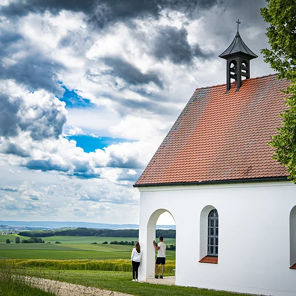 St. Antoniuskapelle bei Belzheim Kleine weiße Kapelle mit rotem Dach in ländlicher Umgebung, zwei Personen stehen davor.